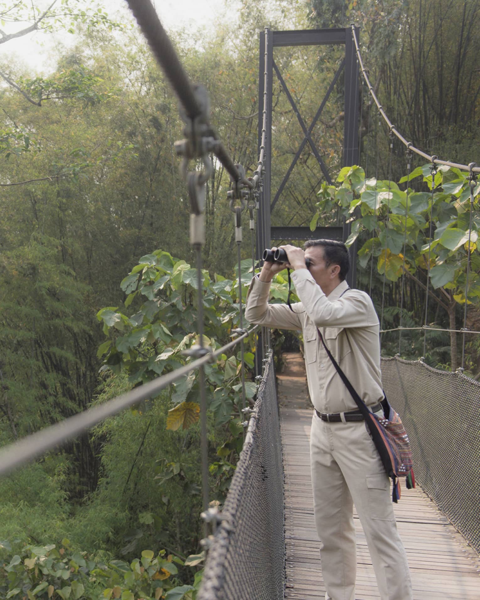 Man wearing artisan bag on suspension bridge surrounded by jungle looking through binoculars 