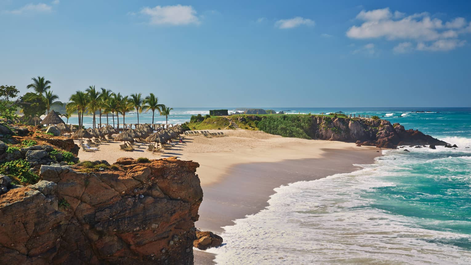 White sand beach lined with lounge chairs, palms, turquoise tides from oceanside cliff