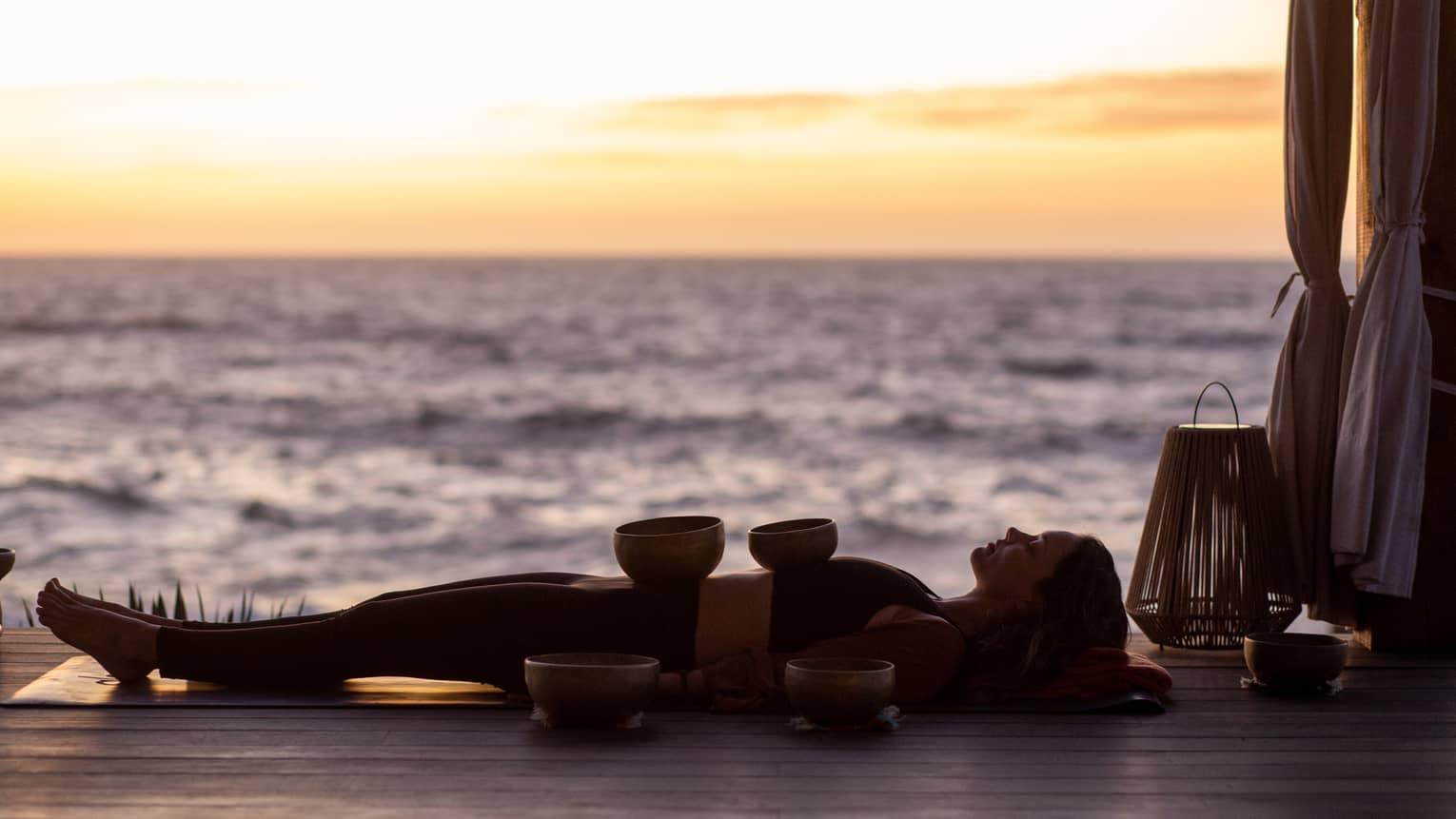 By the sea, silhouetted by a yellow-streaked sky, a person reclined on a yoga mat balances two bowls on their torso.