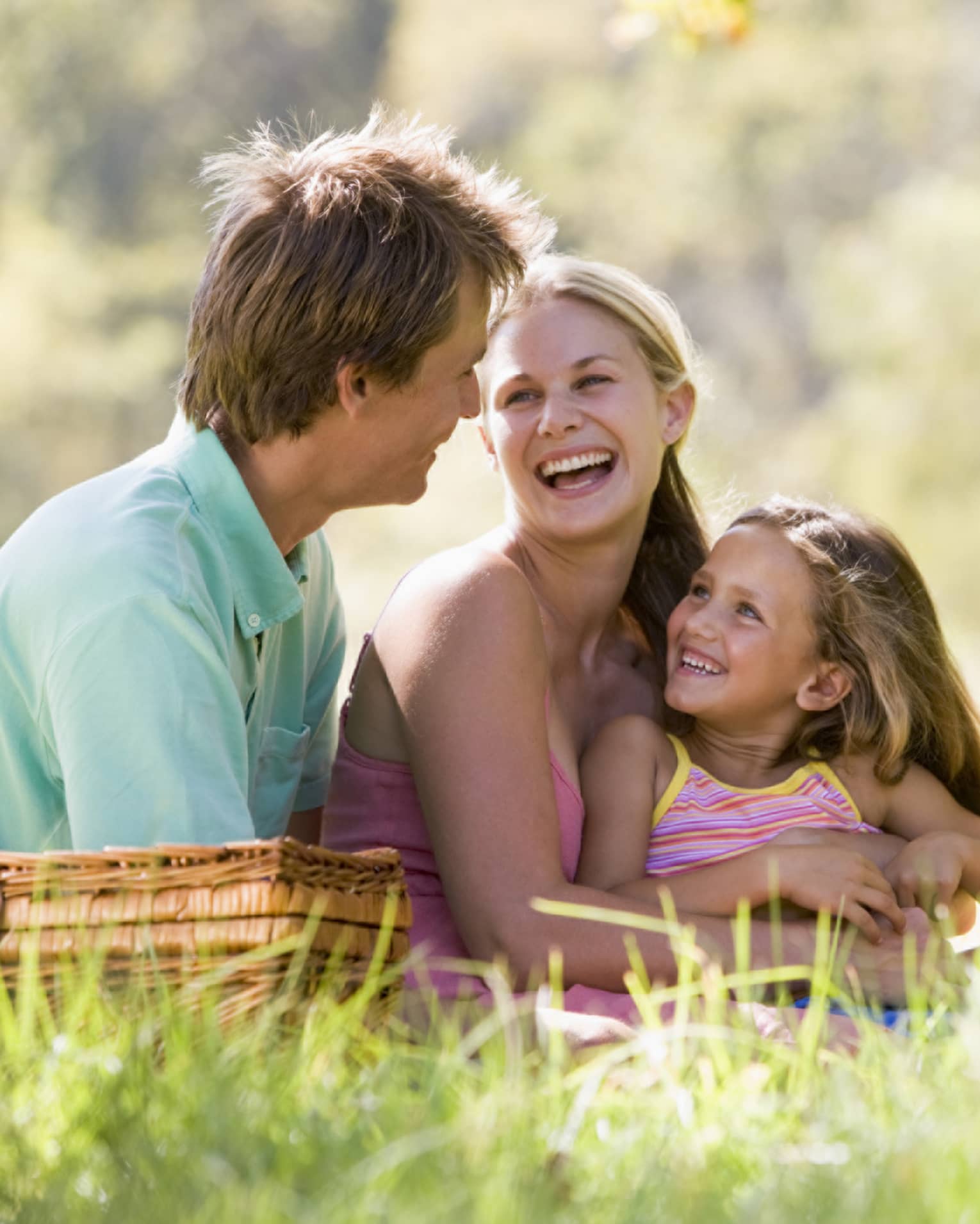 Smiling man, woman and young girl sit in tall grass by picnic basket on sunny day