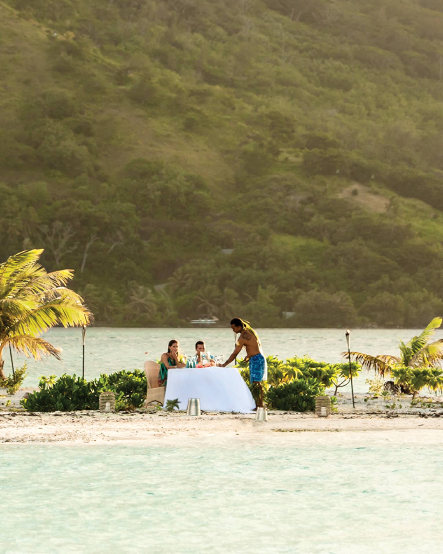 Server stands in beside couple at formal dining table on private white sand motu, lagoon and mountain behind