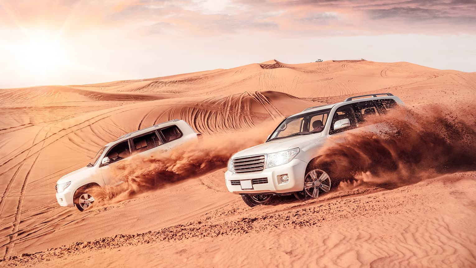 Sand flies from tires of two white SUV's driving across dunes