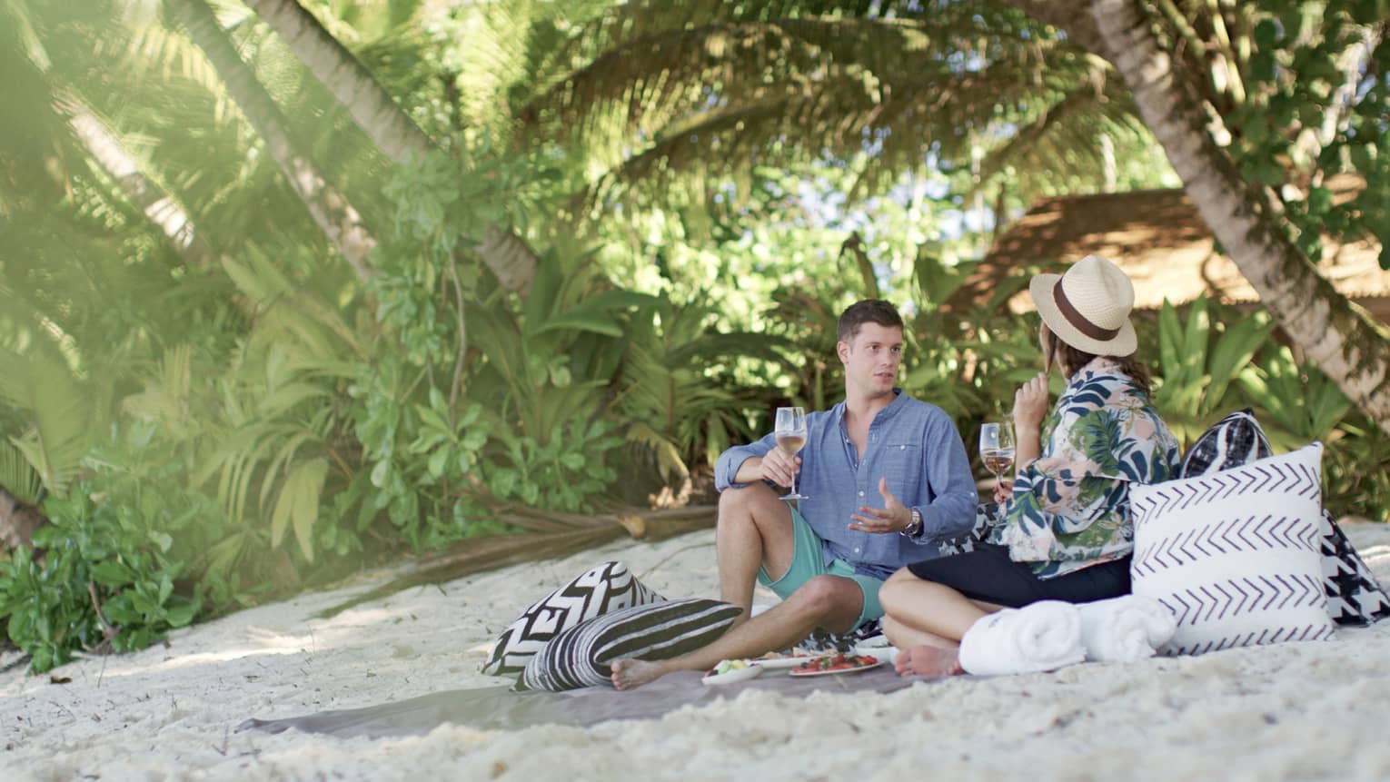 Couple enjoy glass of wine, snacks on pillows on white sand beach