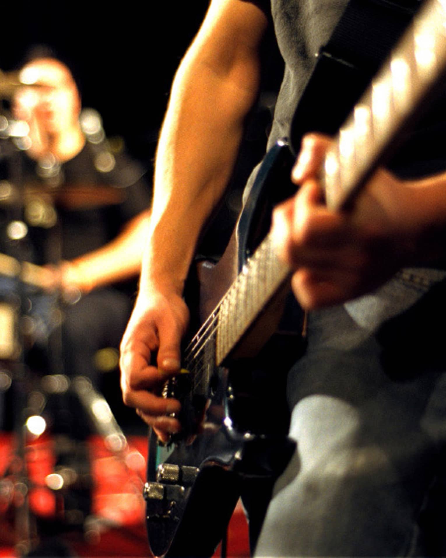 Close-up of musician's arms and hands playing an electric guitar with drumset in the background.