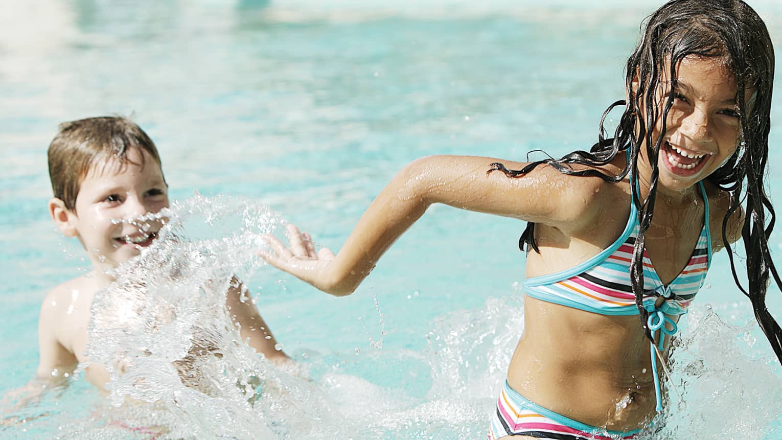 Laughing girl in swimsuit plays in swimming pool, splashes smiling boy