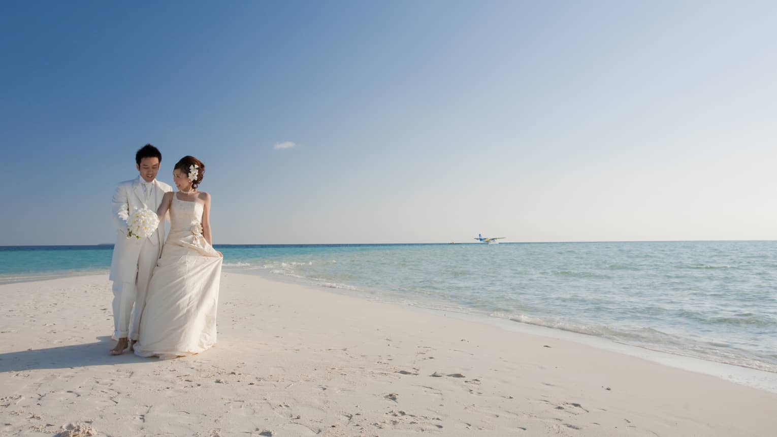 Bride and groom wearing white walk along sandbar by ocean