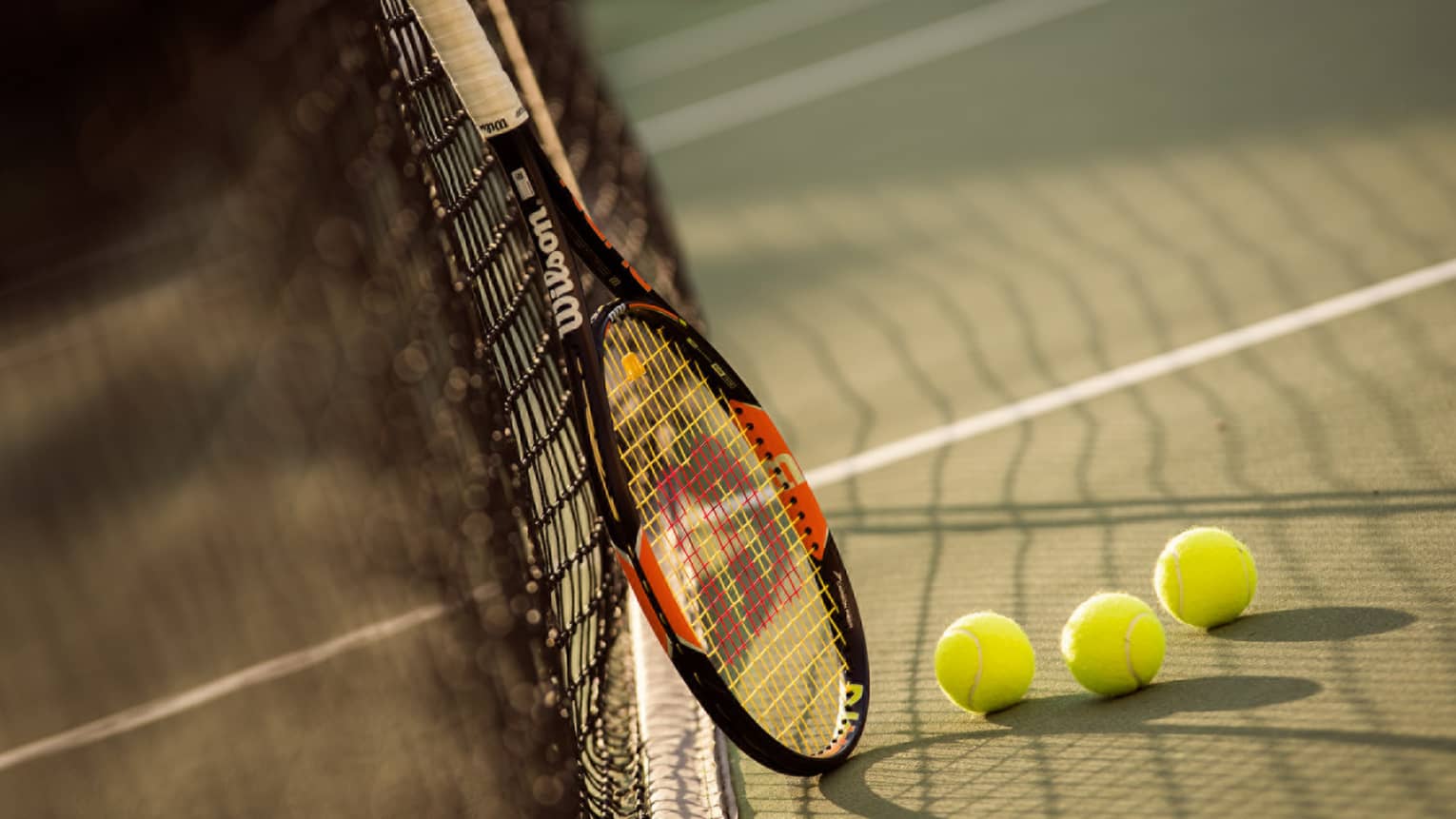 Red and black tennis racket leans against net by green tennis balls on court