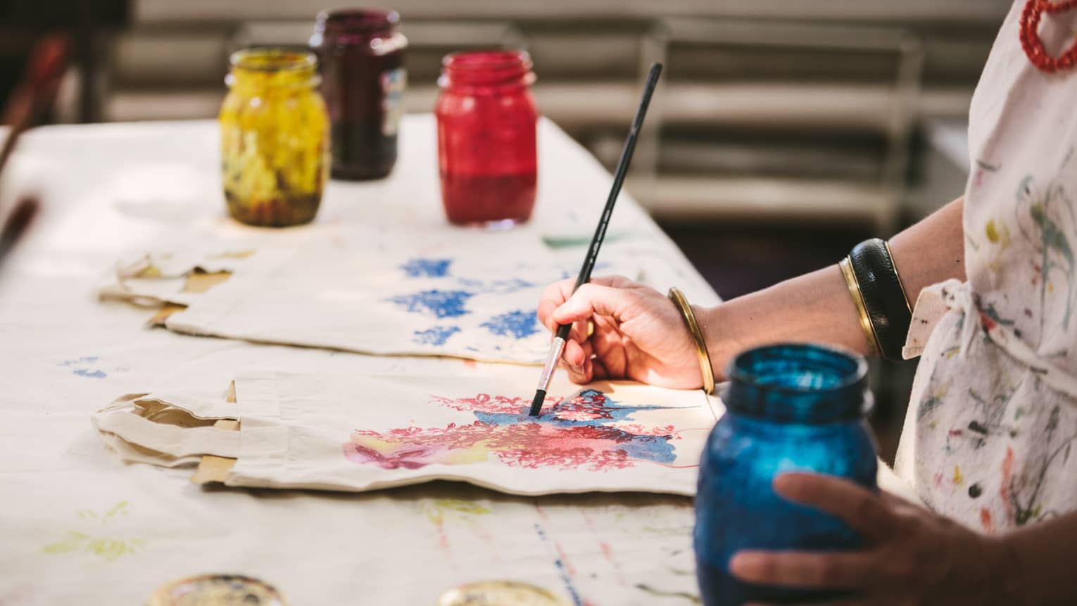 An artist using mason jars filled with red, blue, yellow and black paint to paint images of flowers on canvas tote bags.