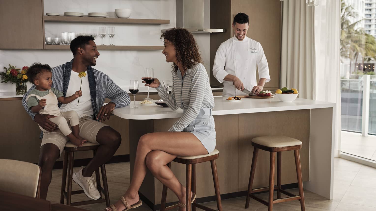 Parents sit at a kitchen island, toddler in dad's arms, laughing over glasses of red wine while a chef prepares a meal. 