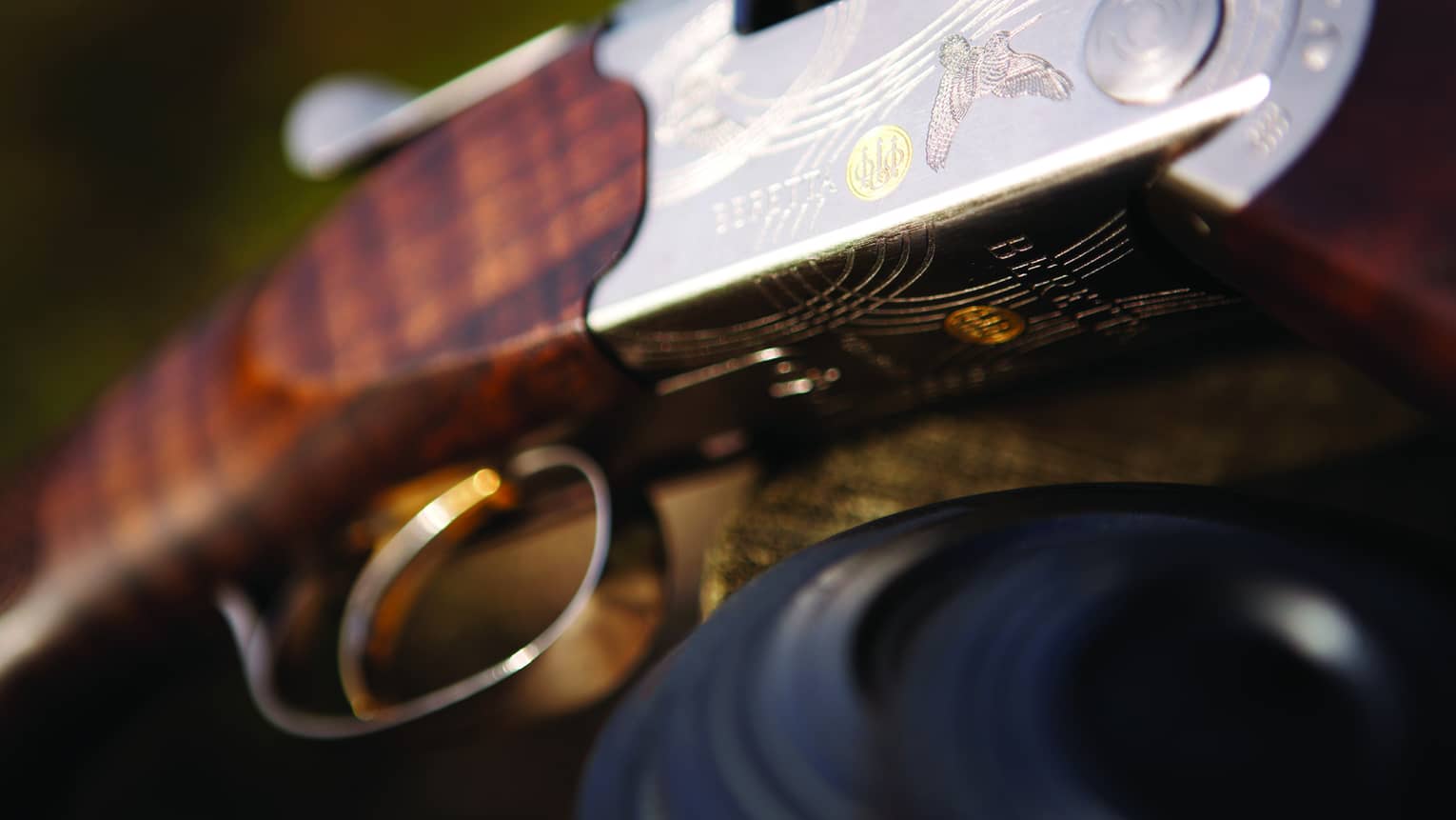 Close-up of trigger and wooden barrel of a rifle with silver engraving of a grouse, the Beretta name, swirls and a gold logo.