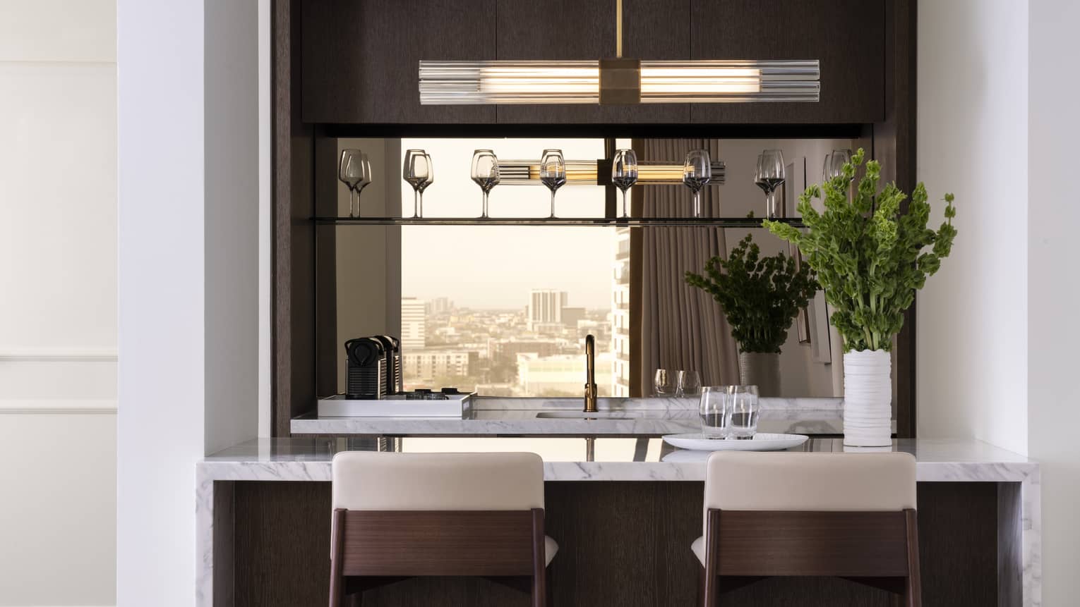 A kitchen with wine glasses on a shelf, herbs, two bar stools, an island and sink.