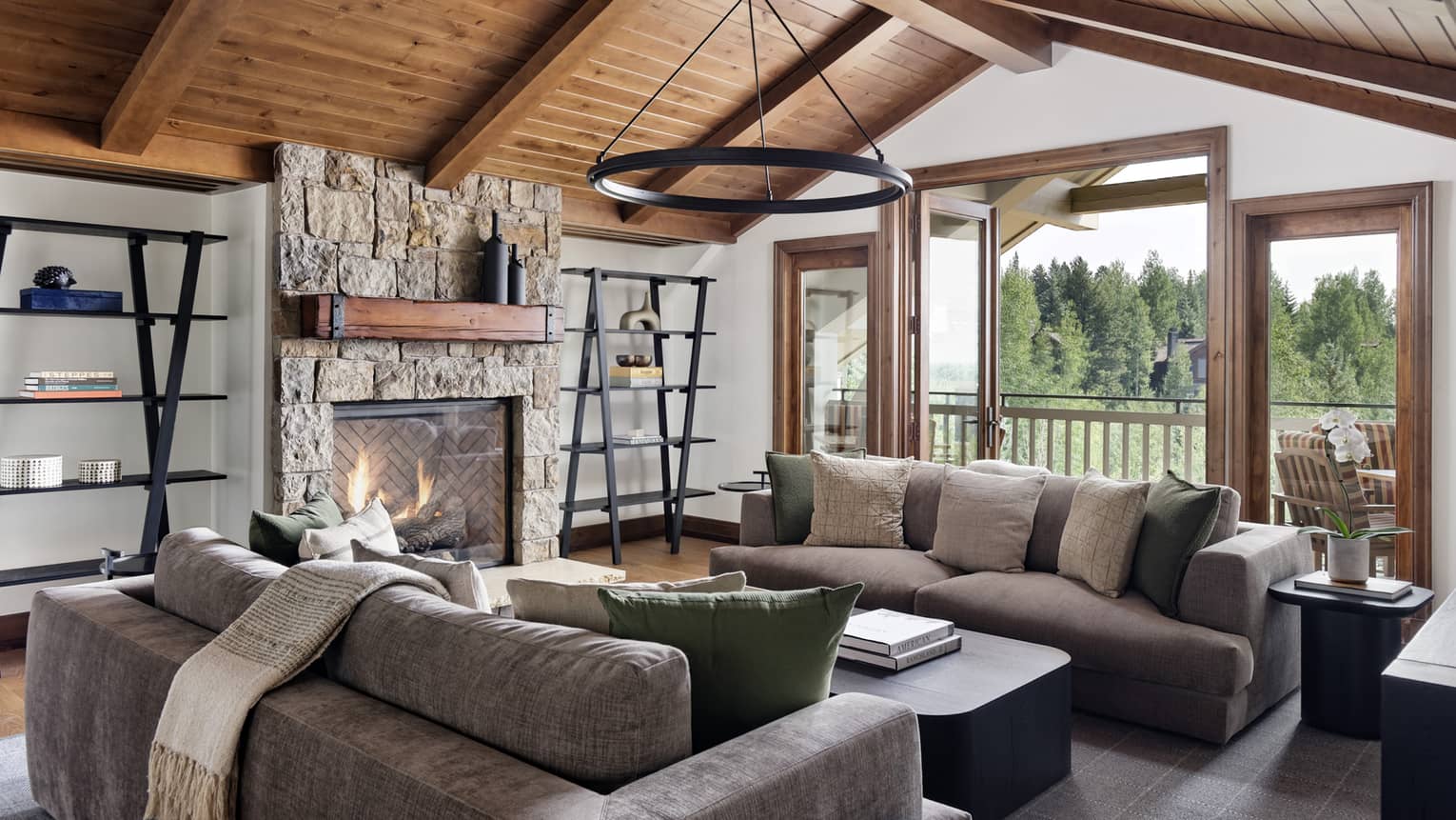 Granite residence living room with vaulted wood ceiling, stone fireplace, grey sofas and a circular chandelier, opening to a balcony with forest view