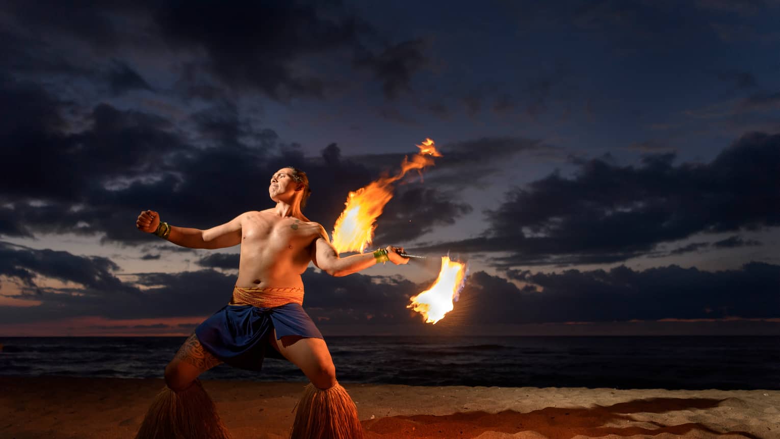 Man wearing traditional Polynesian outfit dances with torch on beach at night