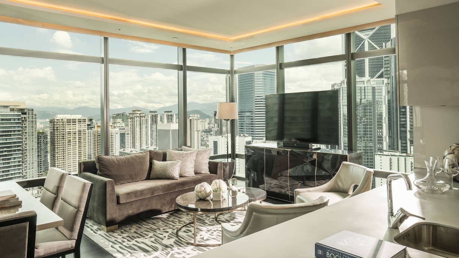 Suite seating area with grey velvet sofa, large TV by corner windows with city views