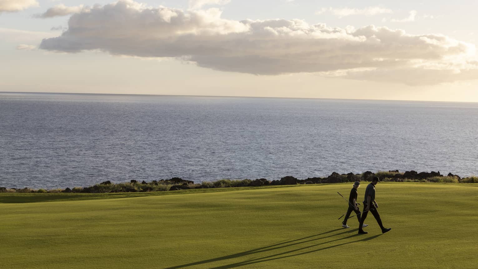 Two people with golf clubs, long shadows reaching out behind, walk a golf course set against calm ocean and fluffy clouds. 