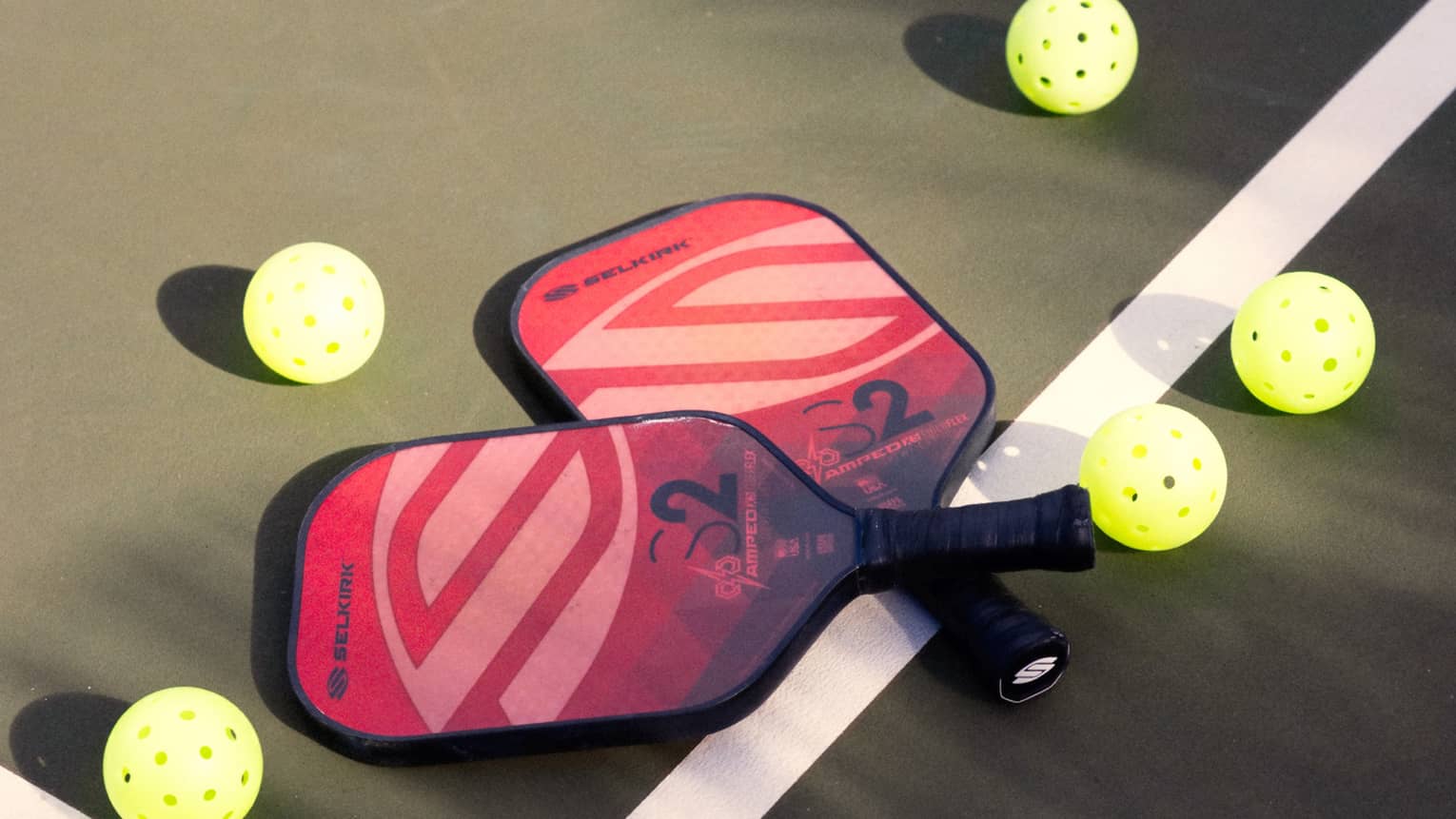 Close-up of two red and pink pickleball racquets, handles crisscrossed on a white court line, surrounded by five pickleballs.