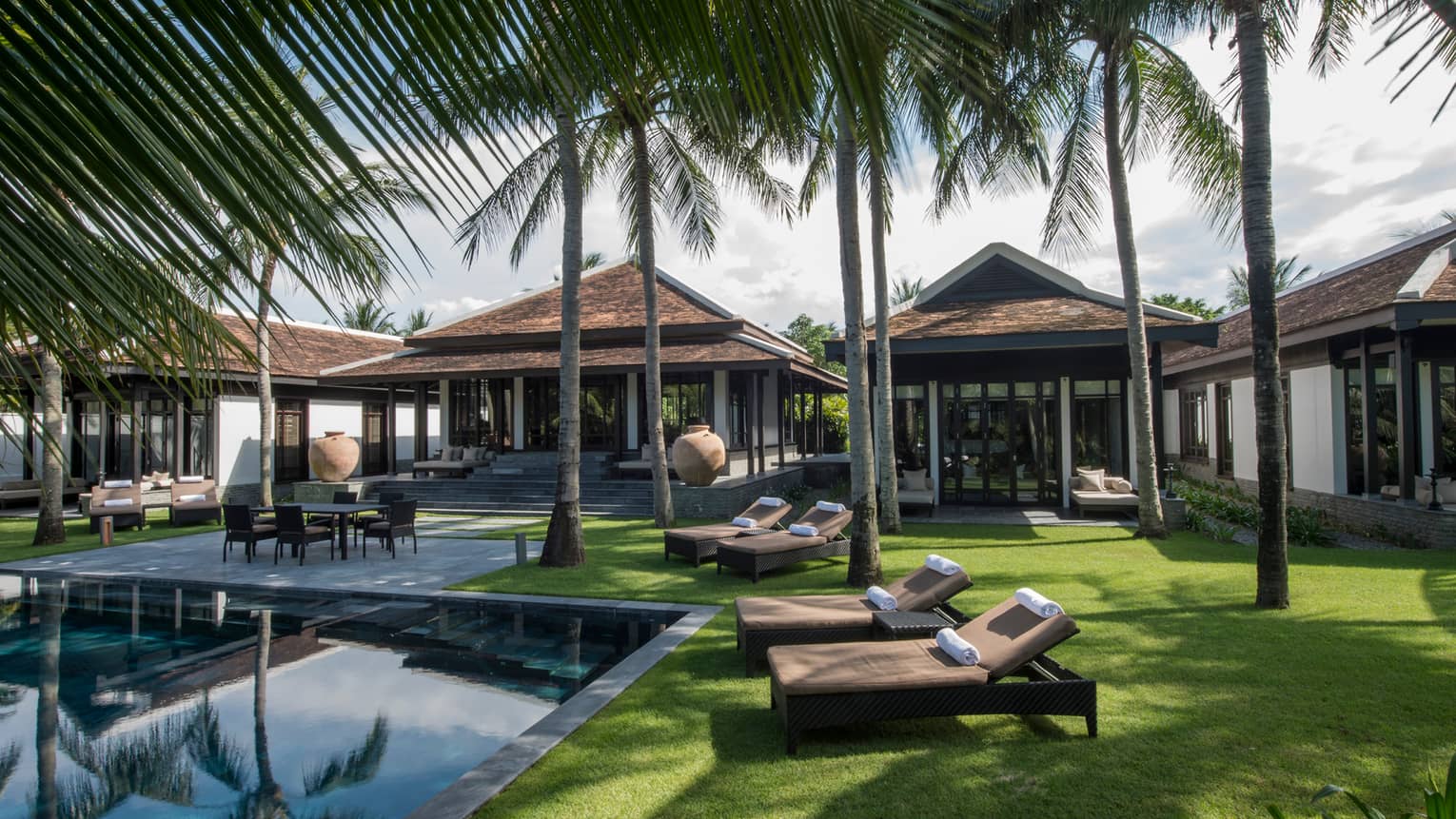 Luxury villa with multiple buildings, surrounded by palm trees, featuring a private pool, lounge chairs and a grassy yard.