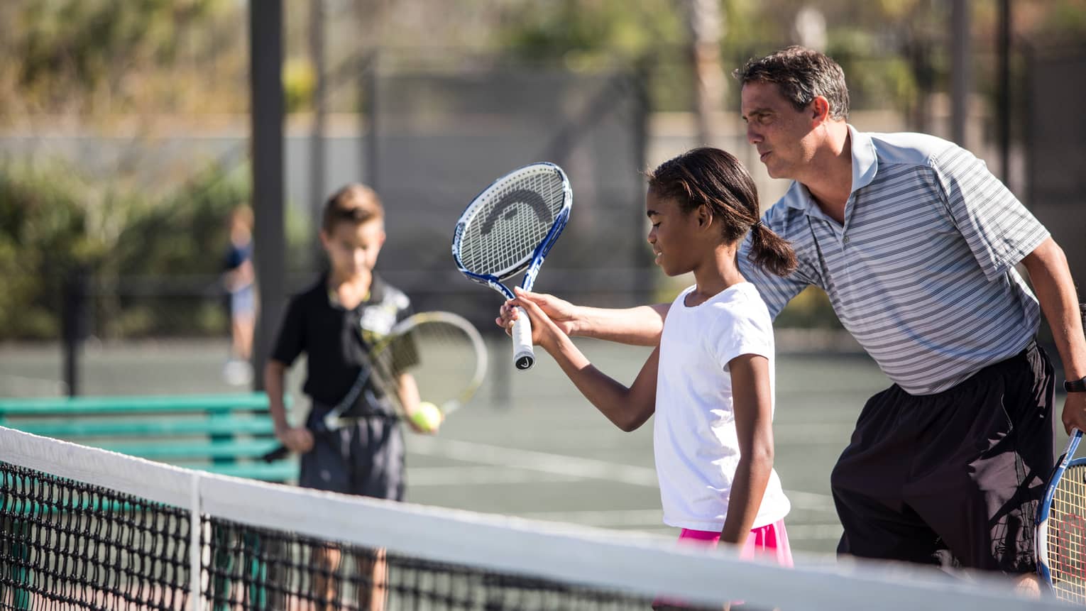 Tennis coach helps young girl hold tennis racket by net as young boy watches