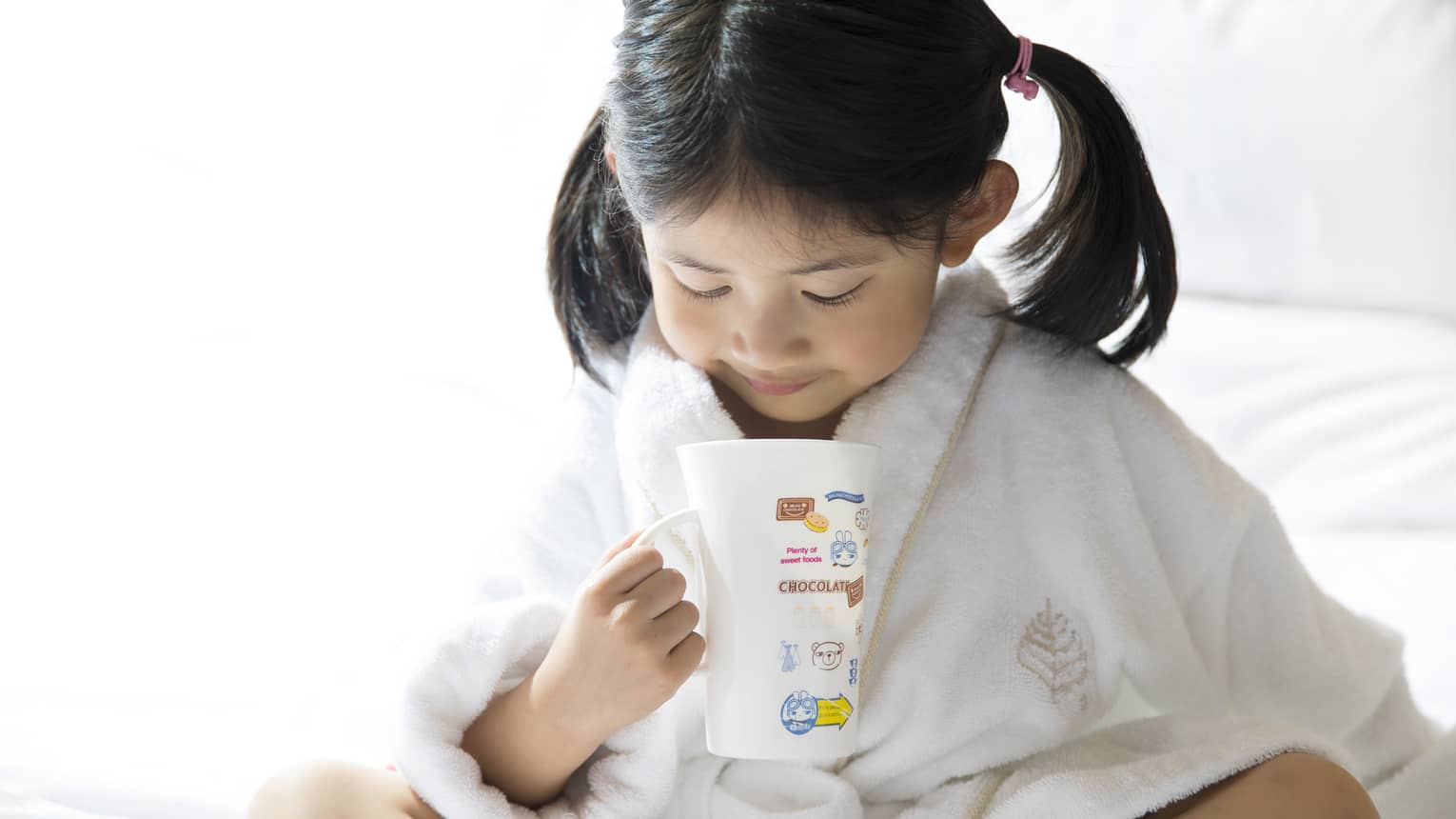 Young child in white bathrobe with hair in ponytails sits cross-legged on bed, holds mug of hot chocolate