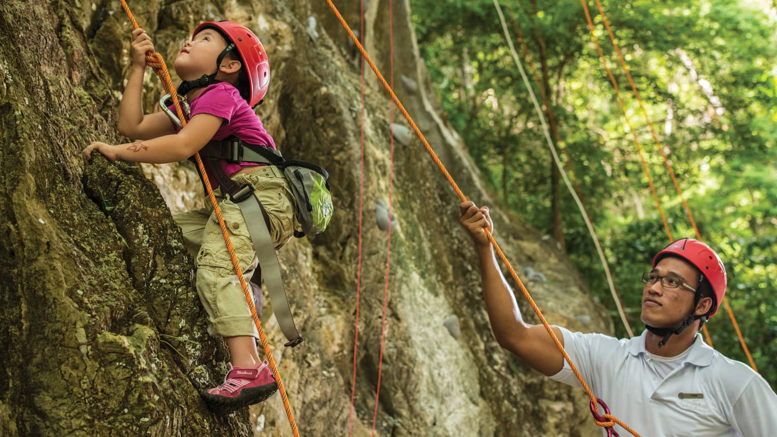 Man holds rope, helps young girl wearing helmet climb rock face