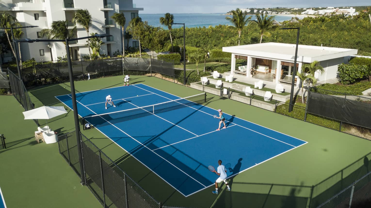 Aerial view of a doubles match on a blue tennis court, with green bordering, palm trees