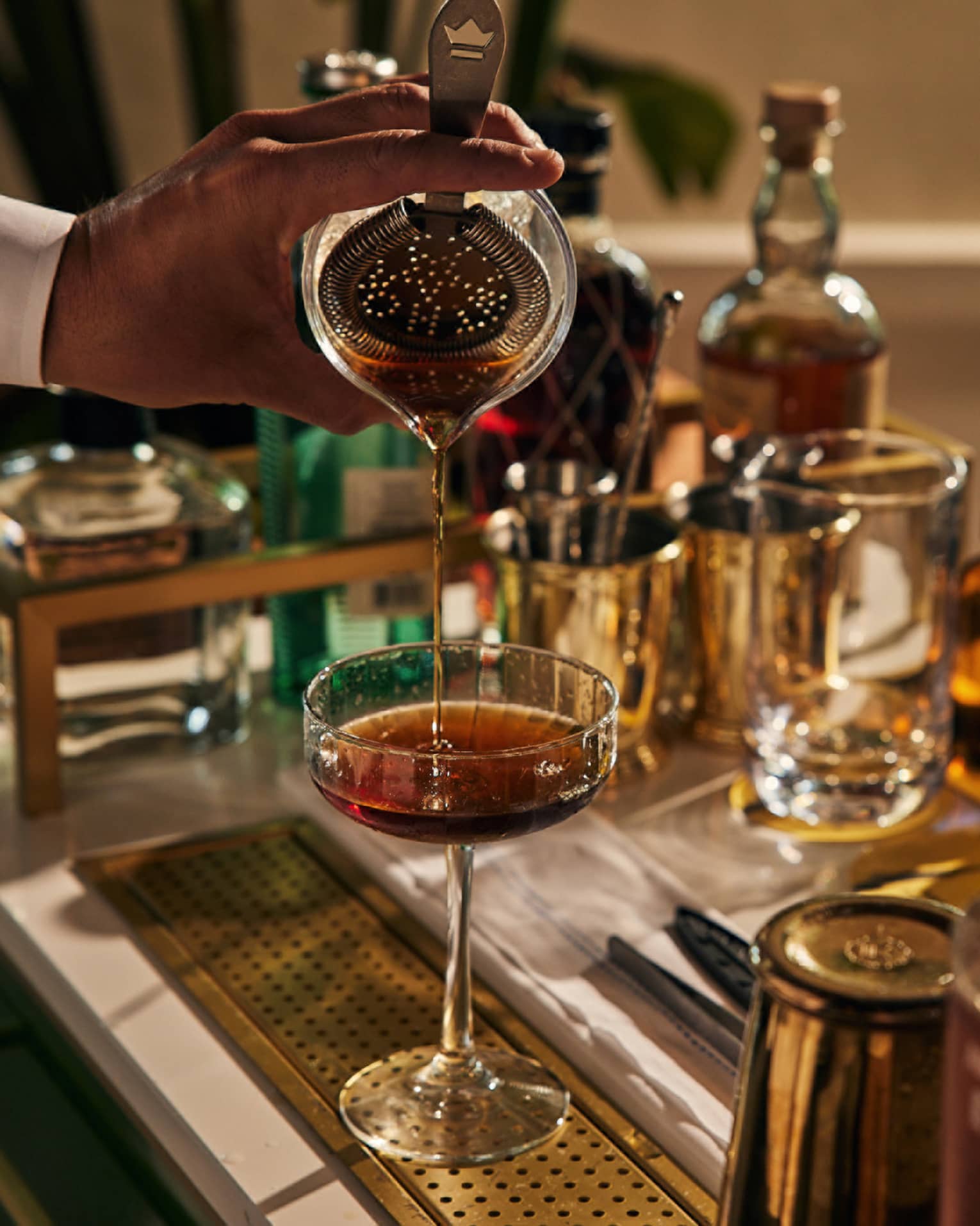 A bartender pouring a drink into a cocktail glass with bottles of liquor around.