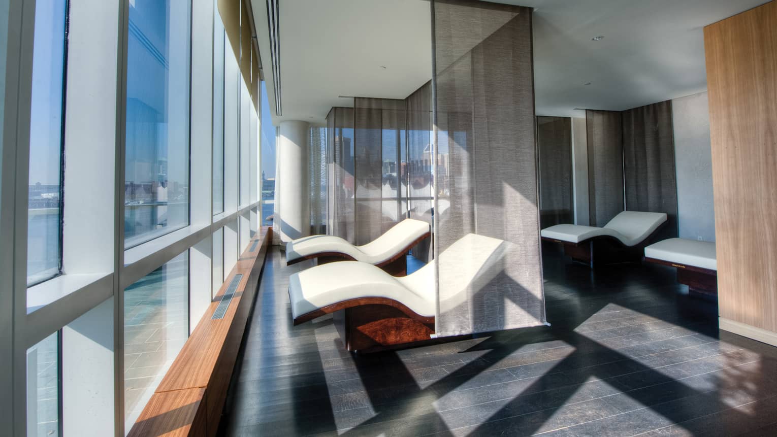Two curved white spa chairs divided by sheer panel curtains in front of floor-to-ceiling windows
