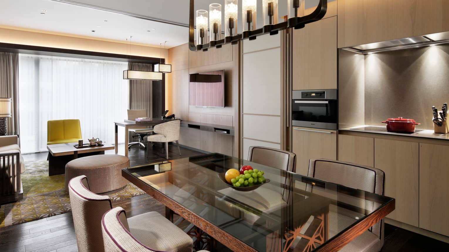 Hotel residence dining table with fruit bowl under long chandelier by modern kitchen cabinets, living room