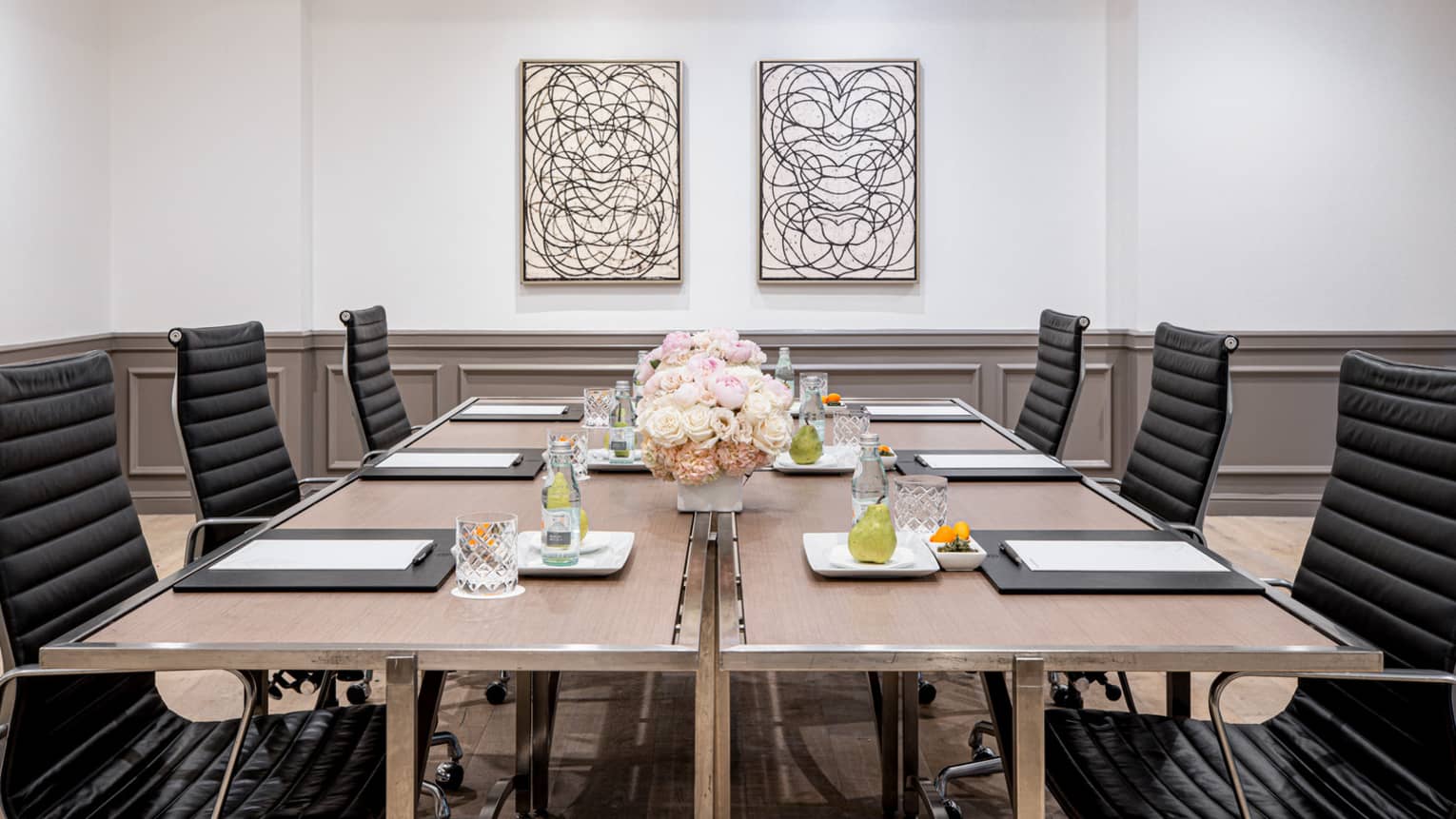 Six black chairs around a wooden table topped with writing pads, beverages and floral arrangements in a bright, spacious conference room