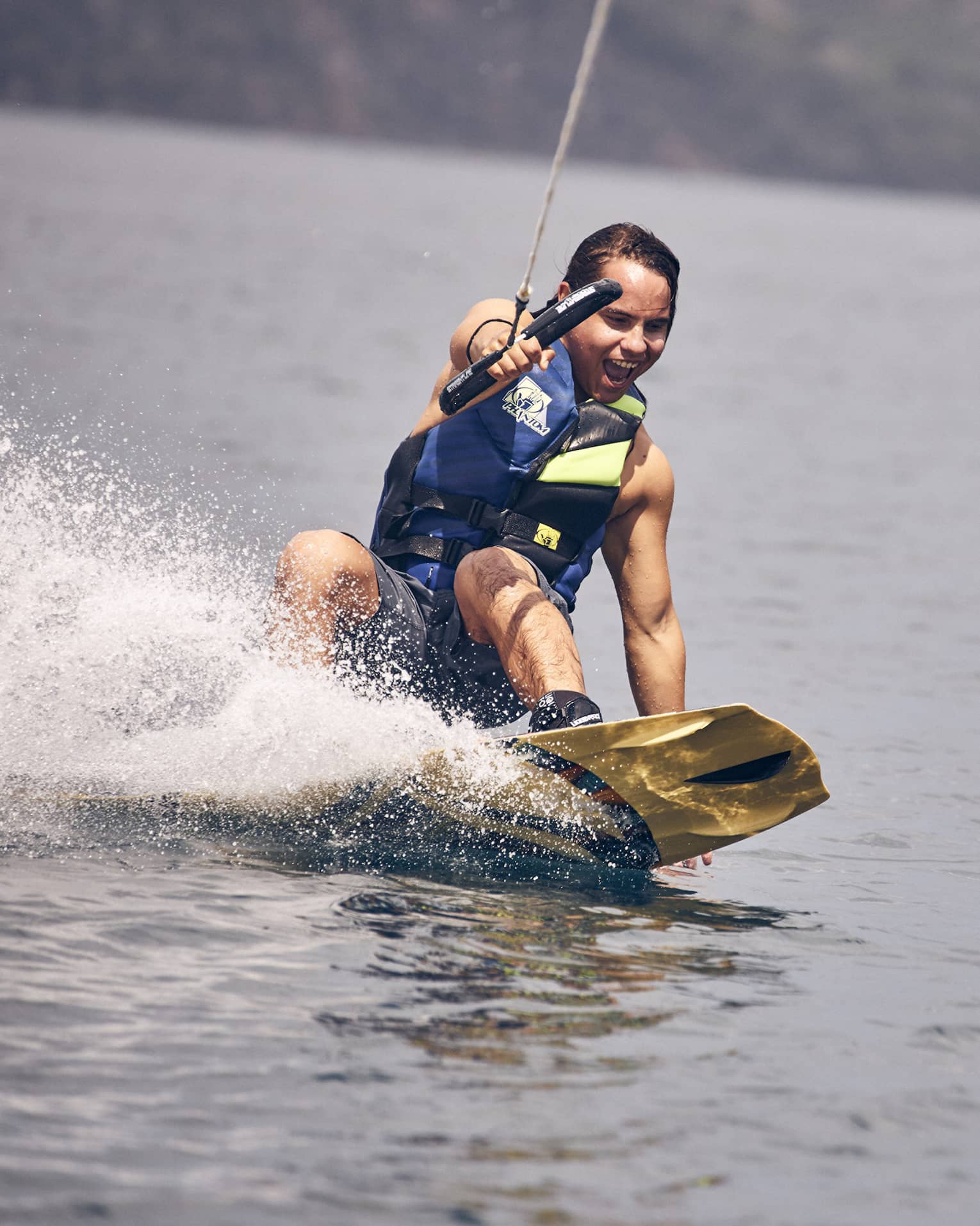 Young man wearing a blue and green life vest and blue swim trunks wakeboarding through the water