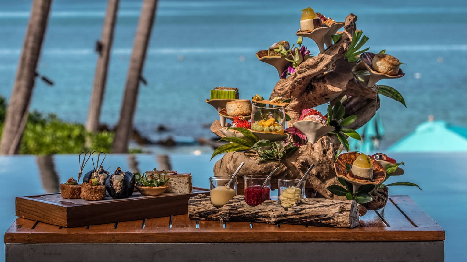 Colourful sweets and spreads arranged on sculpted, tiered driftwood on a teak table, ocean view in the background.