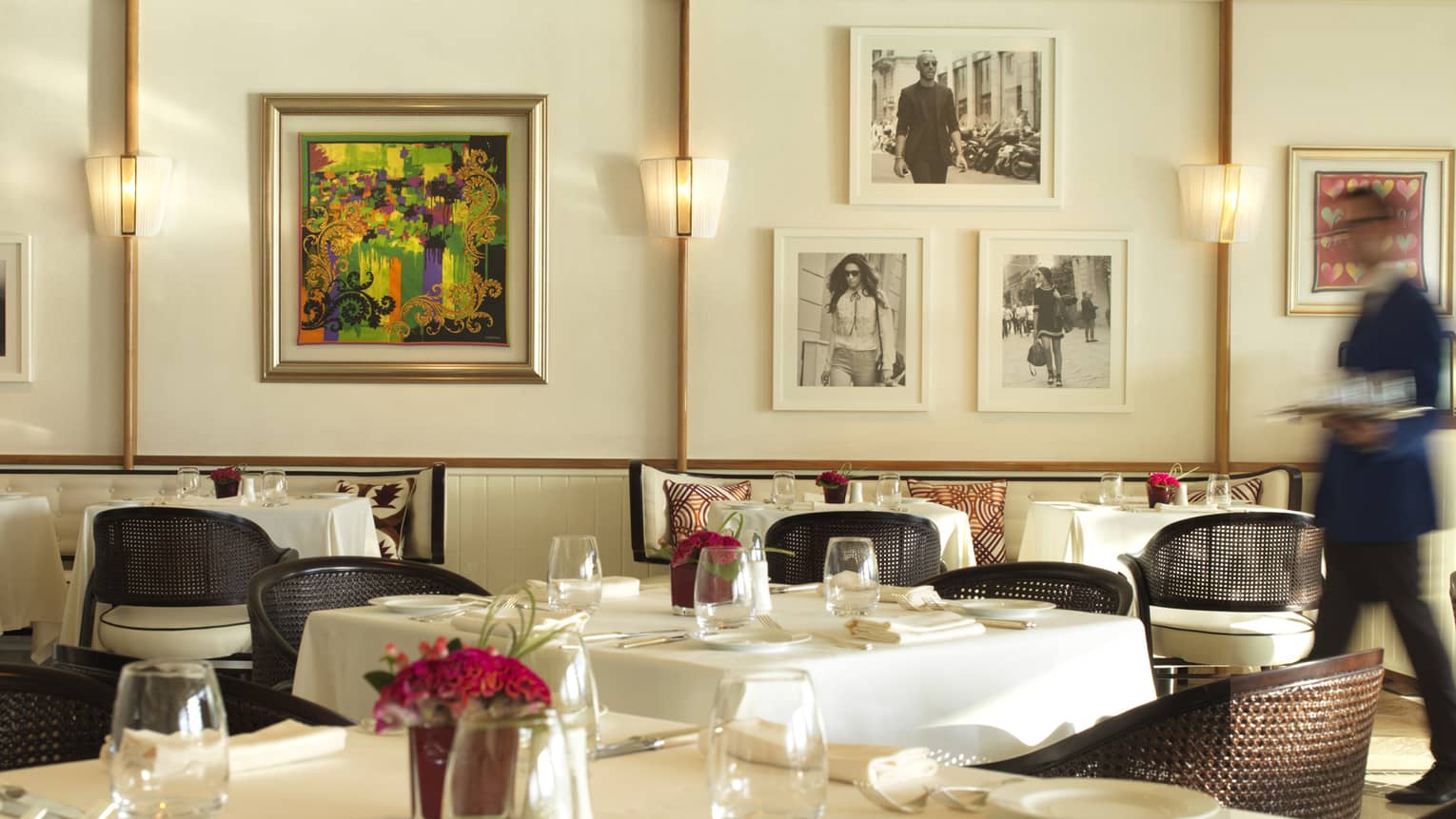 Server in blue suit walks through Cafe Milano dining room with elegant white dining tables, colourful art, framed black-and-white photos