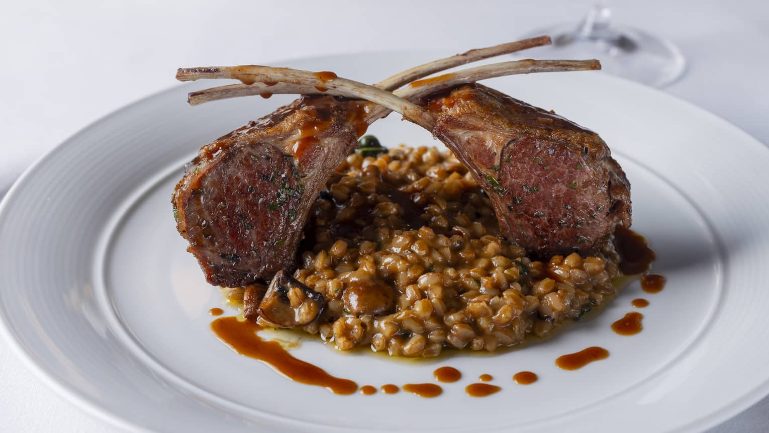 Lamb rack marinated with fresh herbs and lemon, served in a mushroom and spinach farro risotto with red wine and truffle sauce plated on a white, round dish