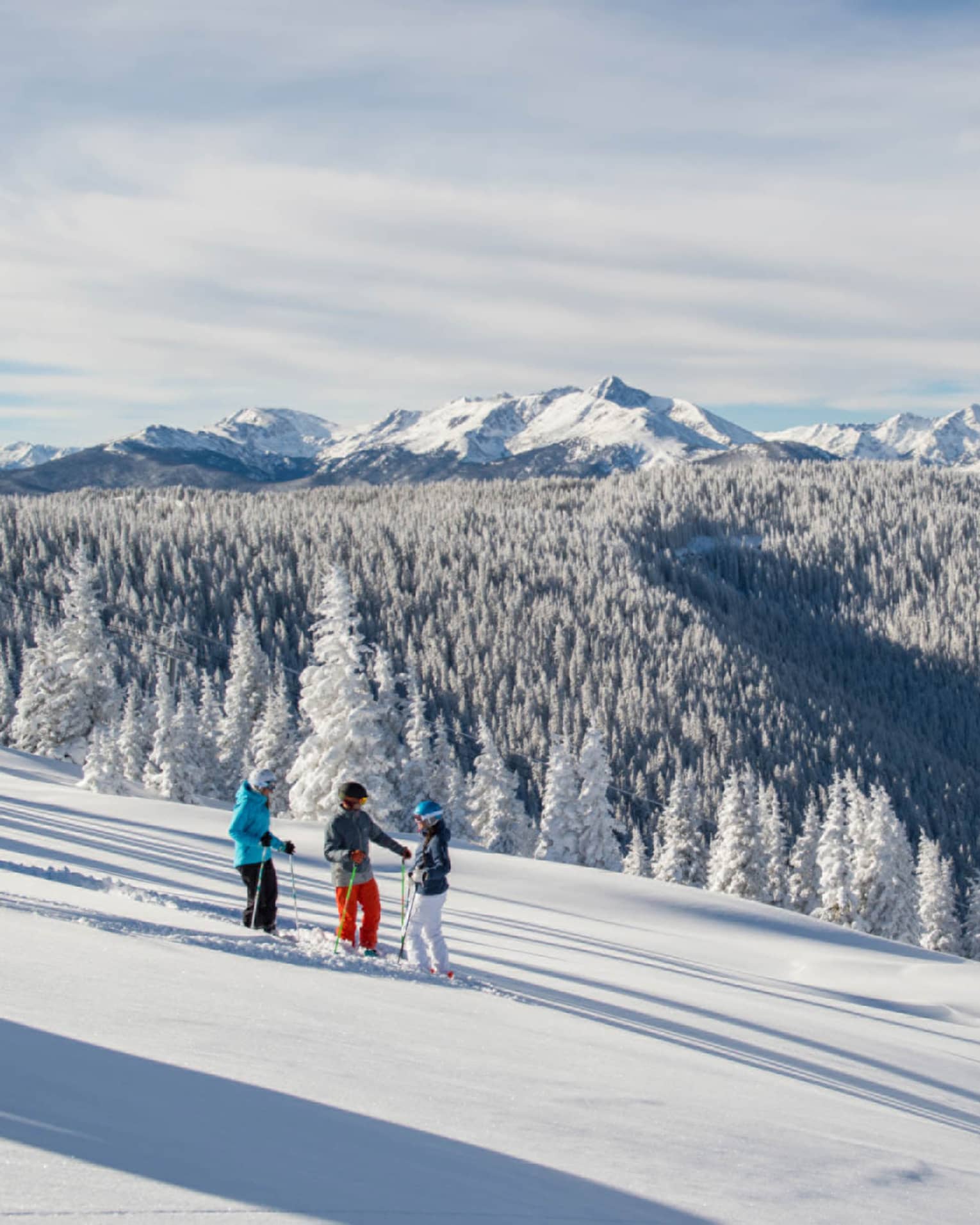 Three guests on skis pause on the ski hill, snow capped trees and mountains in distance 