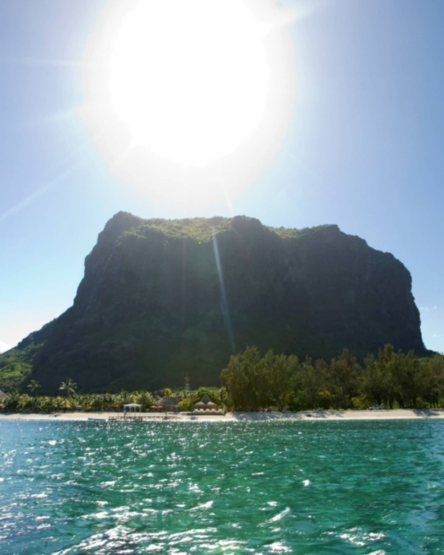 Long view of a forested monolith mountain sitting atop crystal waters. Sun beams down from above in a clear blue sky.