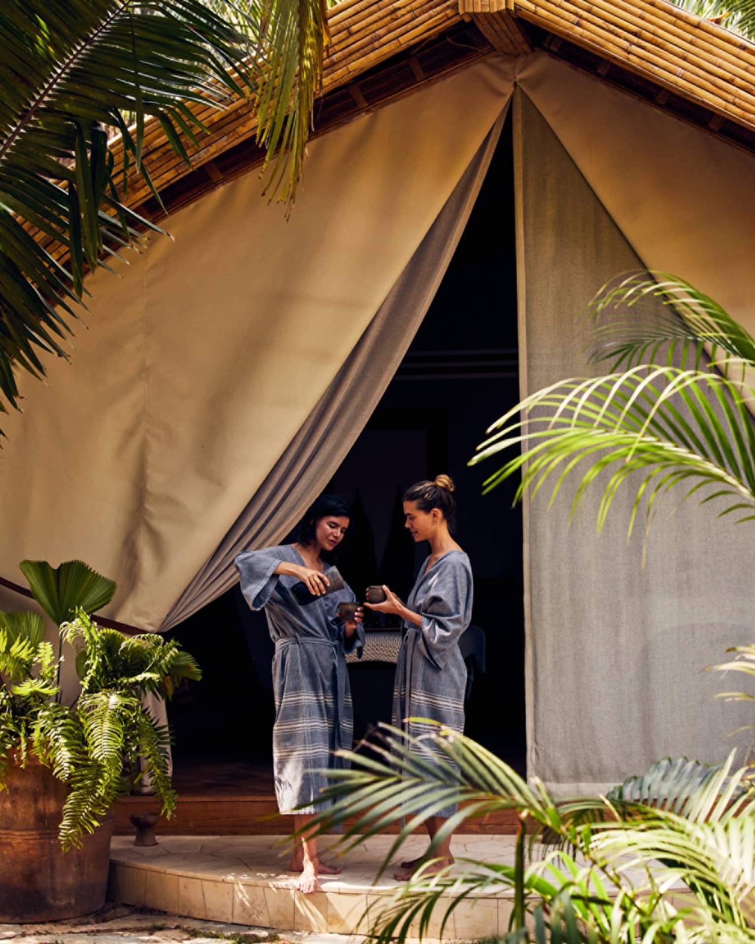 Two women in spa robes entering a spa tent surrounded by palm trees.