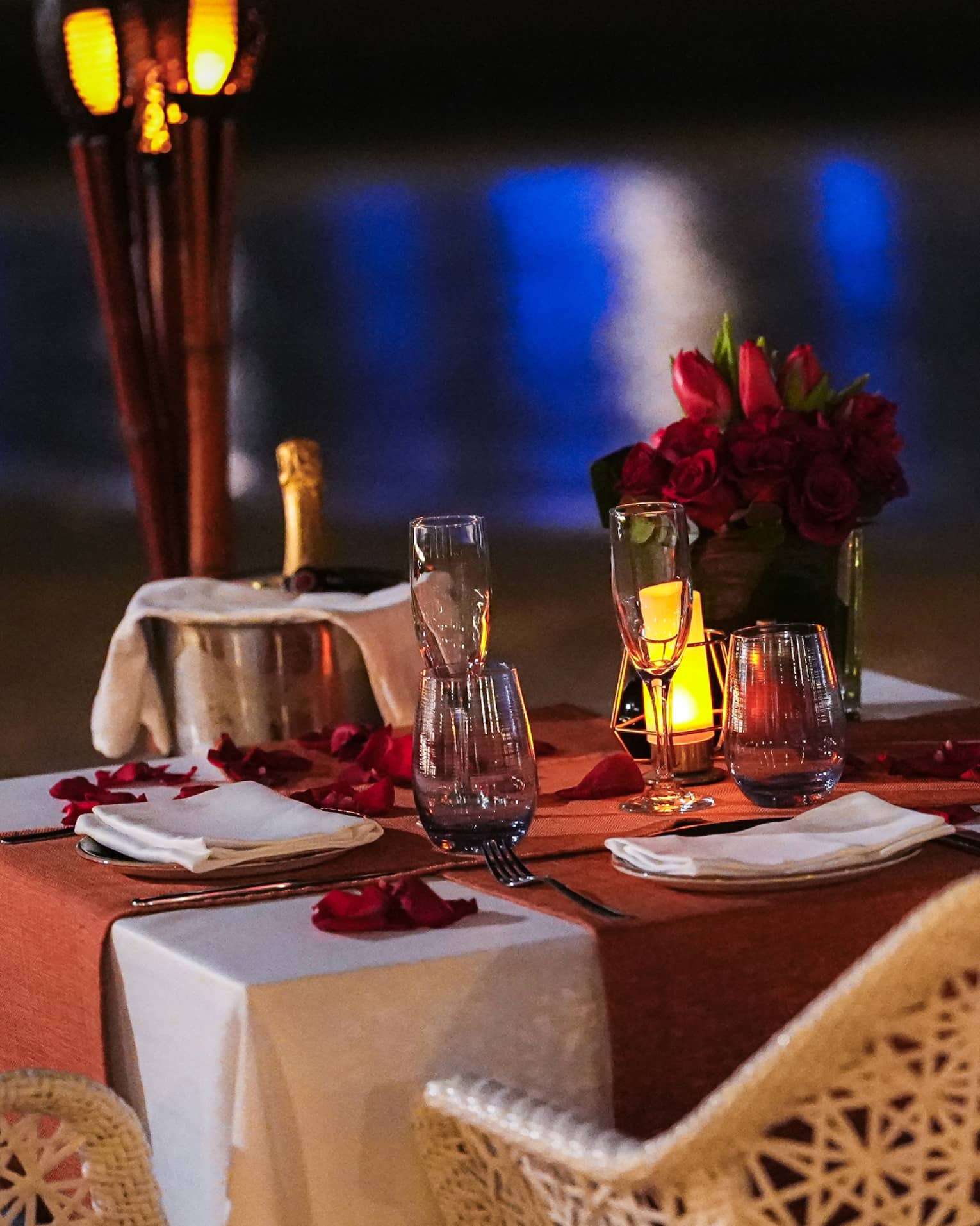 Table for two set with chilling champagne, flutes and a rose bouquet, aside torches and the gleaming reflection on the water.