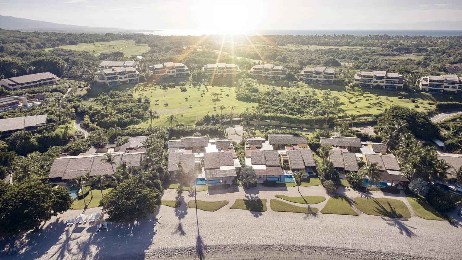 Aerial view of green lawns, trees and estate rooftops behind white sand beach at sunset