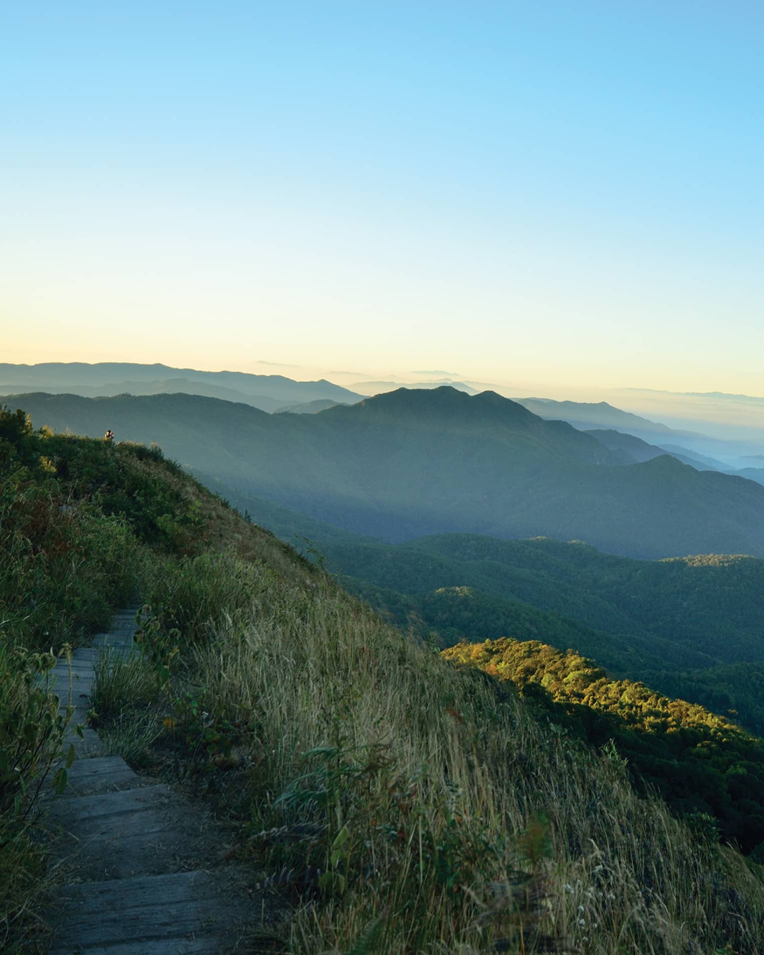 View from an elevated grass-lined path of low, forested mountains receding into the misty distance under blue and orange sky.