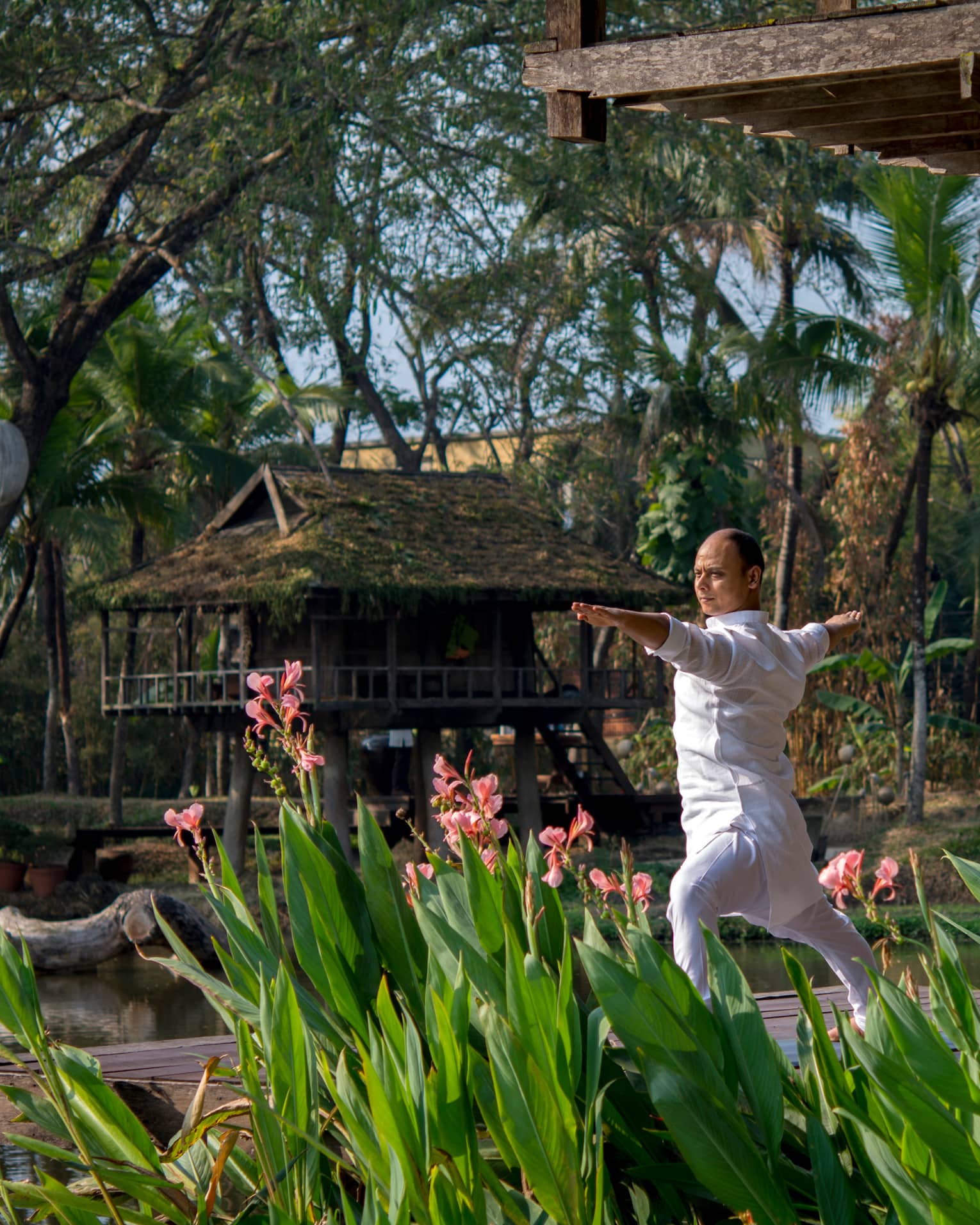 Resident yogi Dheera wearing white in yoga pose with arms outstretched by tall flowers, leaves