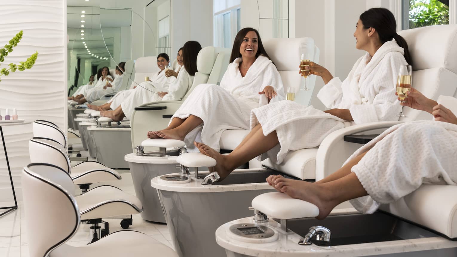 Three women wearing bath robes sit in pedicure spa chairs