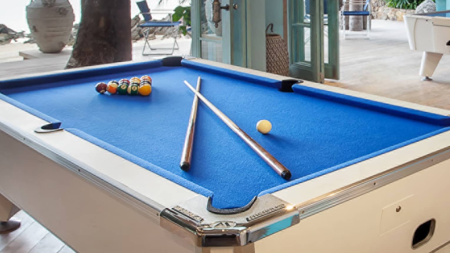 Blue billiards table with pool balls, sticks by seating area, open wall to patio