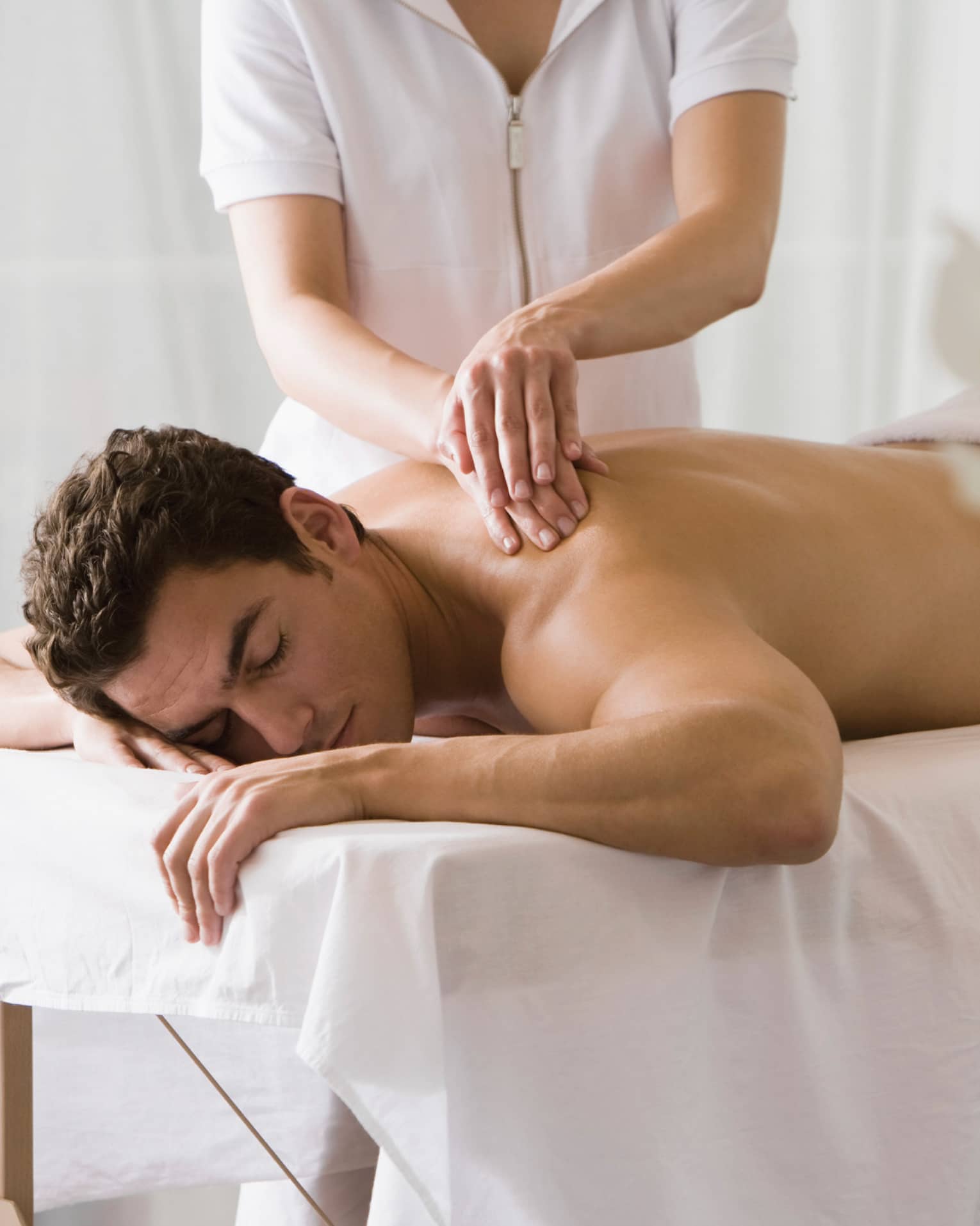 Man lays on white table with eyes closed as masseuse massages his bare shoulders