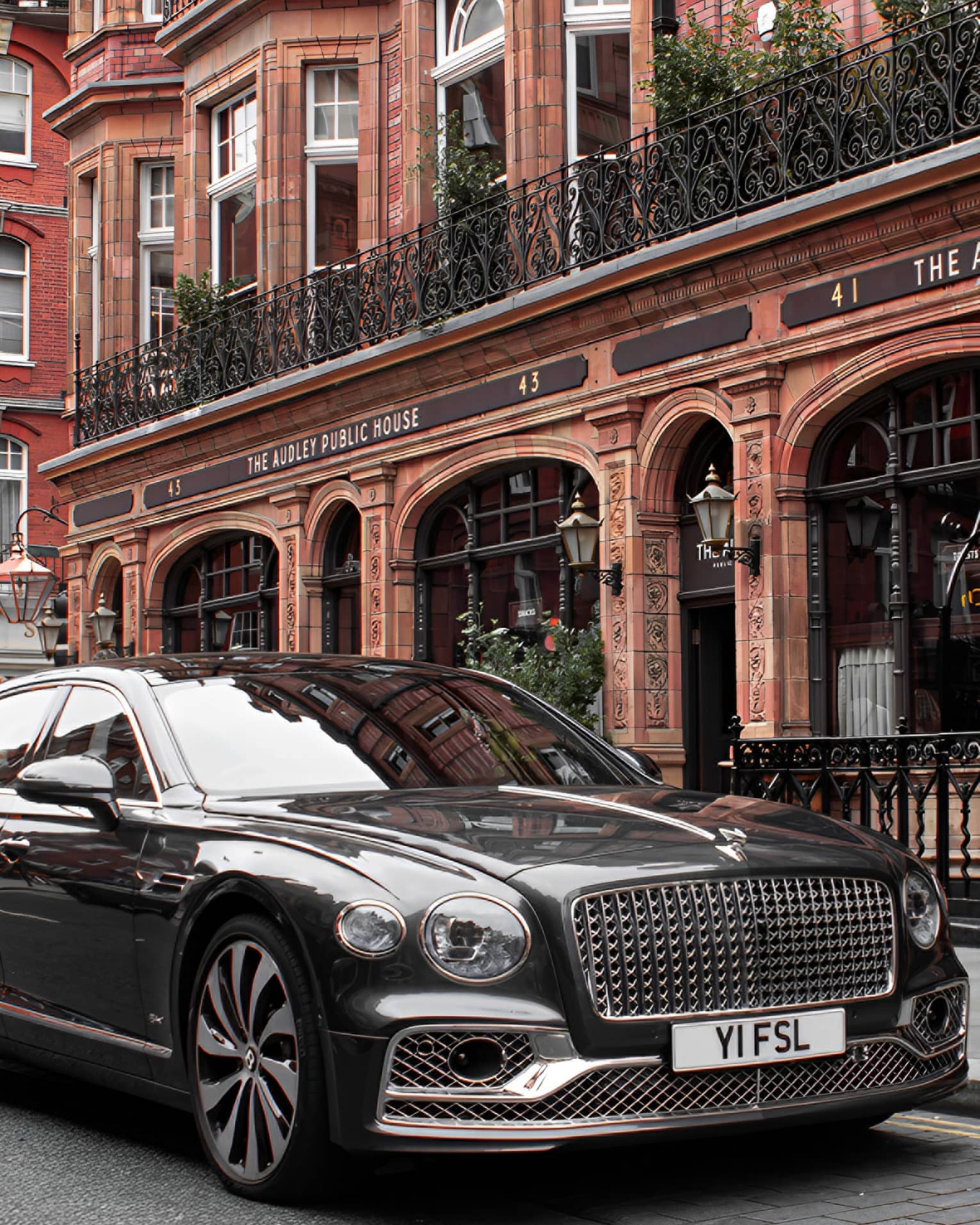 Close-up front view of shiny black modern Bentley with an elaborate grille parked in front of a restored Victorian building.