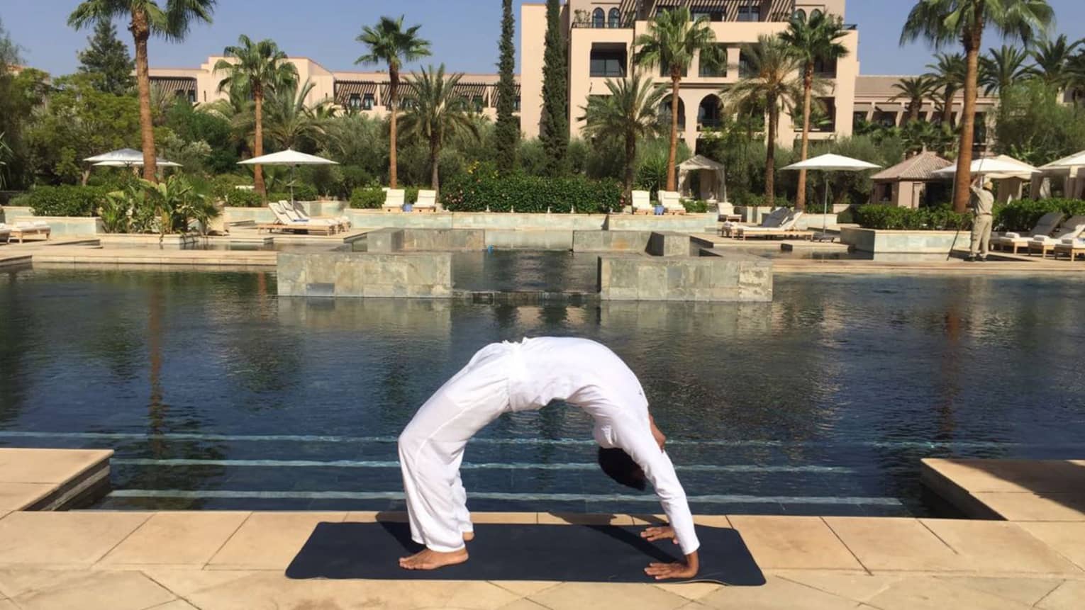 Person wearing white poses in arched yoga bridge pose beside outdoor swimming pool
