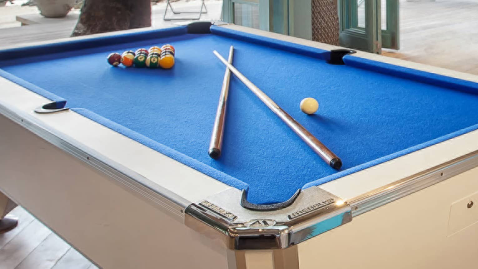 Blue billiards table with pool balls, sticks by seating area, open wall to patio
