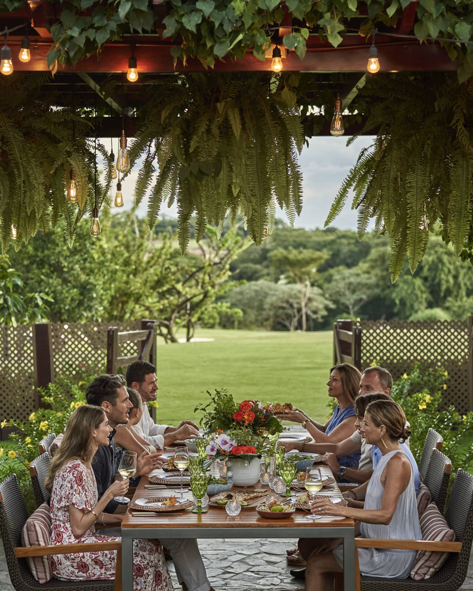 Eight people sit around a long dining table set up outside beneath a canopy of greenery and string lights