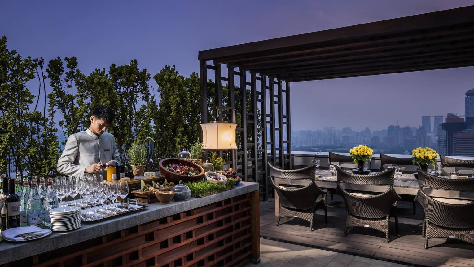 Bartender prepares drinks, cheeses, salads at long Imperial Suite rooftop bar by private dining table at night
