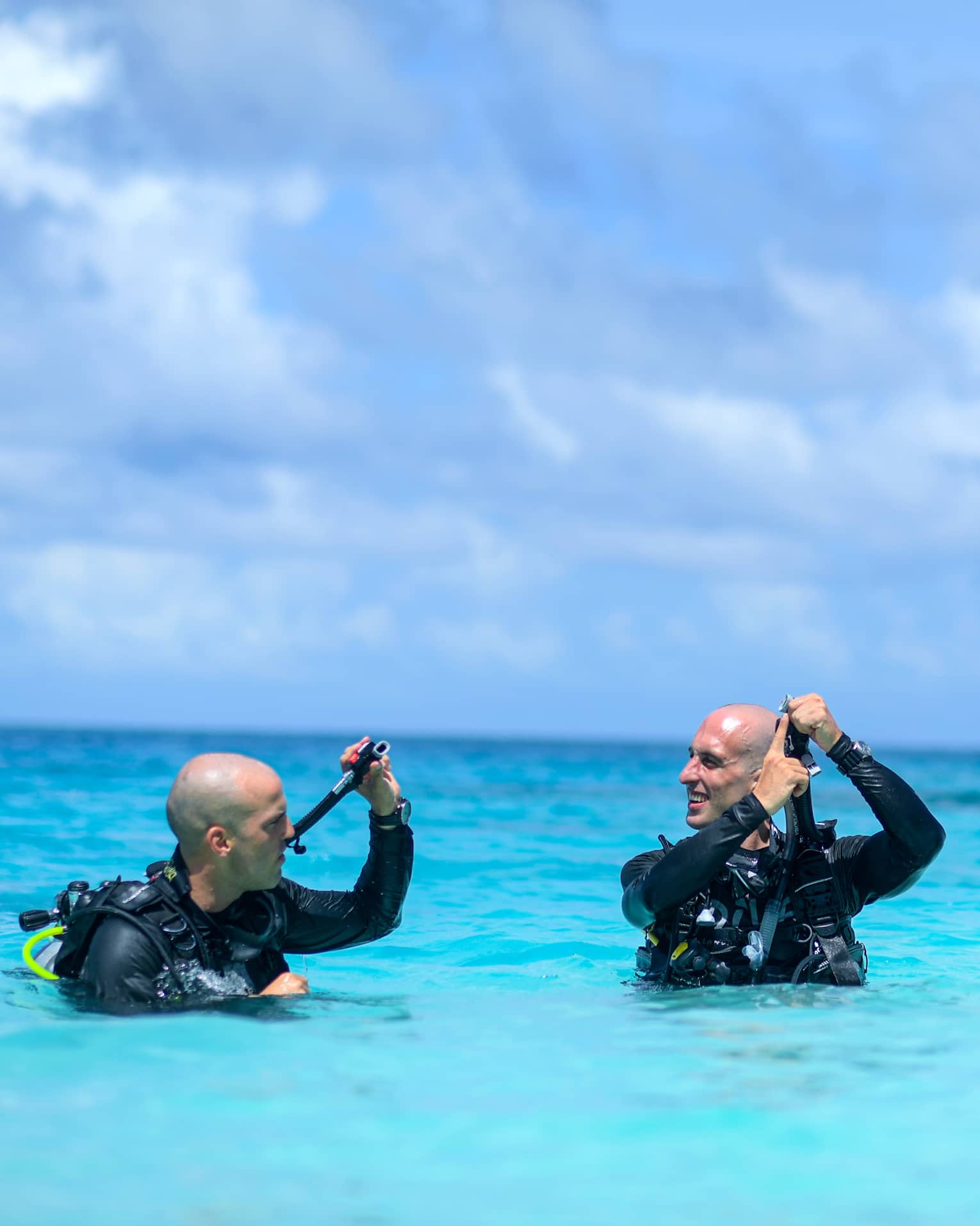 Scuba instructor showing mouthpiece to guest as they stand wearing scuba gear, chest-deep in azure water under fluffy clouds.