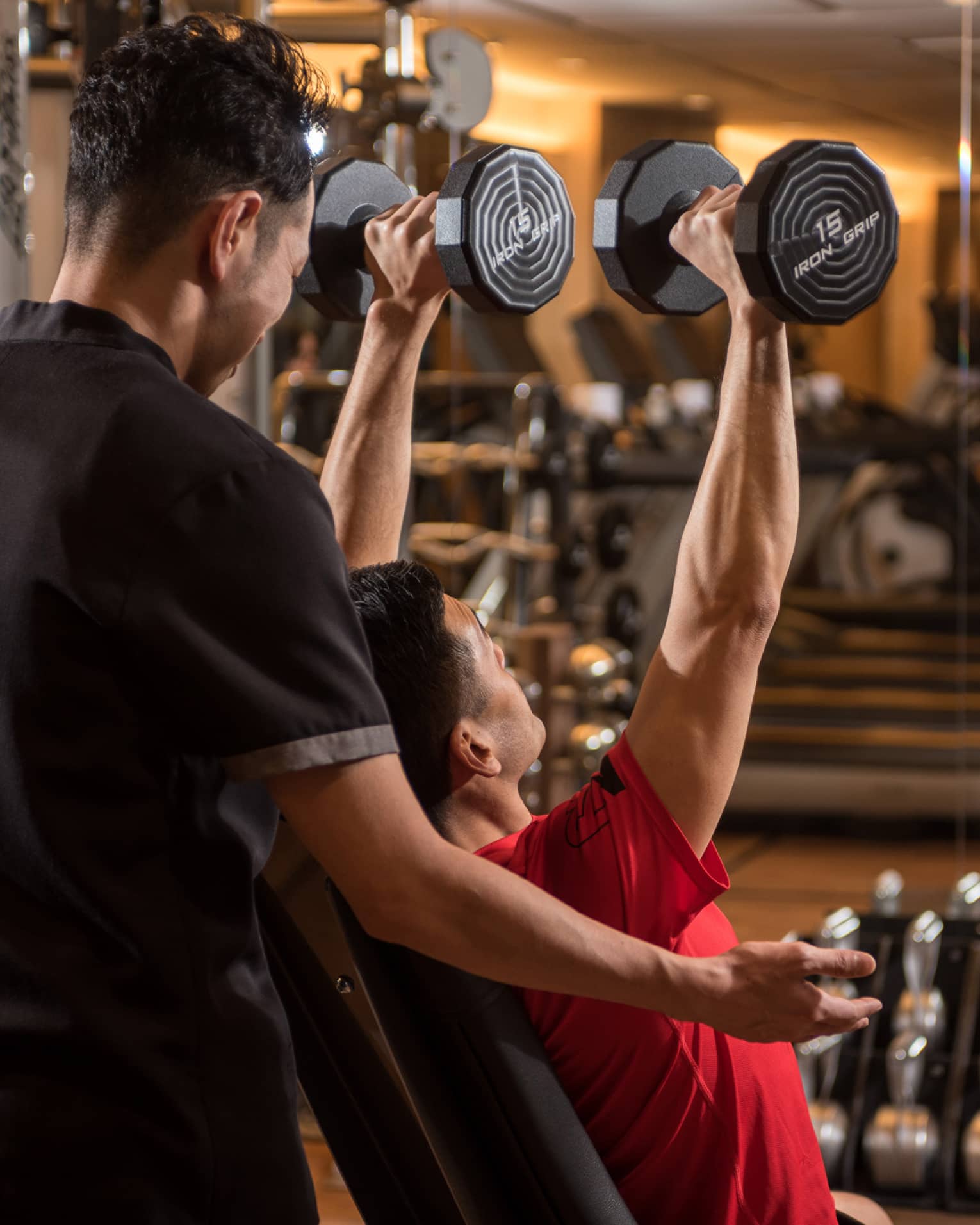 Man in chair in front of mirror lifts two 15 lb. weights above his head while personal trainer assists